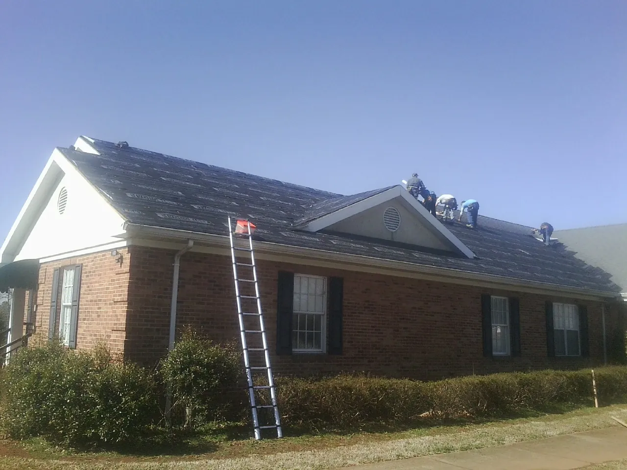 Crew tearing shingles off a brick ranch roof with a ladder set — Emergency Roof Repair work by our crew in Onsted