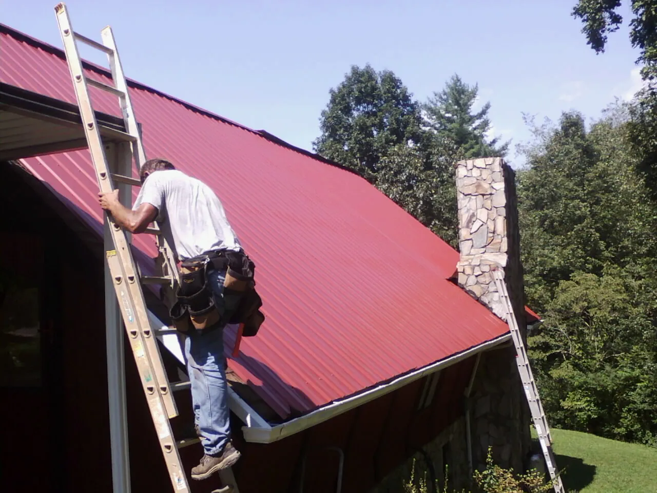Roofer on a ladder beside a red metal roof and stone chimney — Soffit & Fascia Repair by Roofing Brothers in Onsted